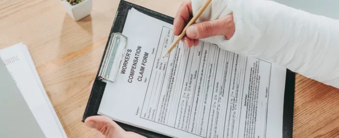 Injured person with bandaged arm filling out workers’ compensation claim form on a desk.