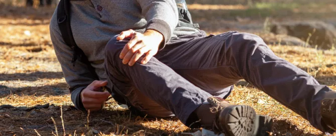 Man in outdoor gear sits on a forest floor, clutching his knee in pain, under sunlight—representing injury and premises liability in West Virginia.
