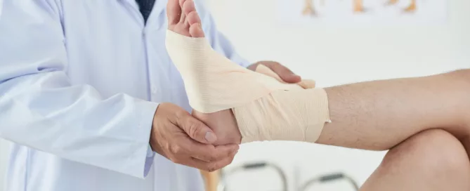 Doctor gently examining a bandaged foot in a clinical setting with anatomical charts and a walker nearby.