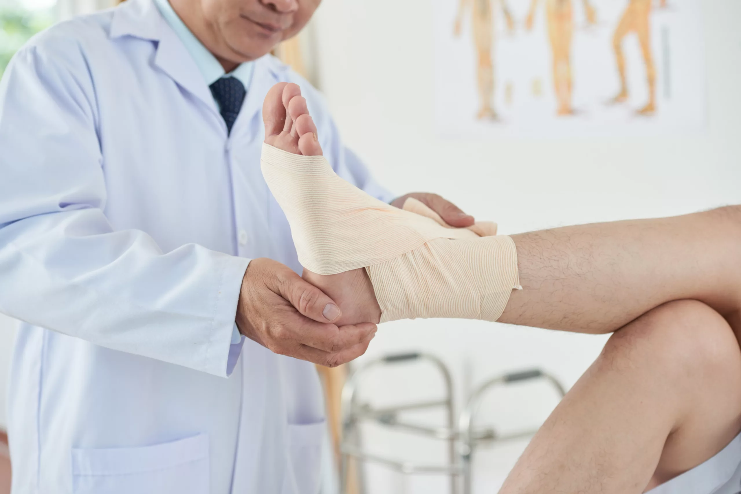 Doctor gently examining a bandaged foot in a clinical setting with anatomical charts and a walker nearby.