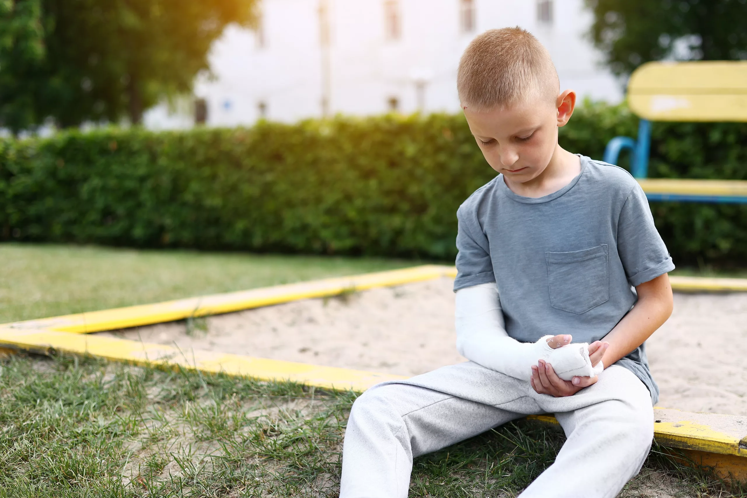 Young boy sitting on grass near a sandbox, looking thoughtfully at his cast-covered arm — conveying childhood injury, concern, and the potential for a minor’s personal injury claim.