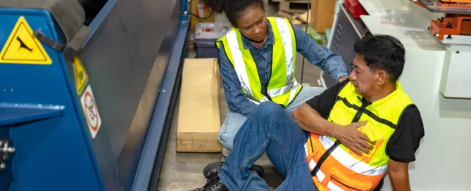 Factory worker in safety vest helping a seated man who holds his chest, surrounded by industrial equipment — an emergency workplace injury scene that conveys concern, urgency and possible legal or compensation issues.